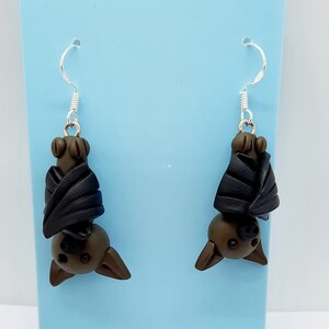 May include: A pair of dangle earrings featuring a brown bat with black wings hanging upside down. The earrings are displayed on a light blue background.