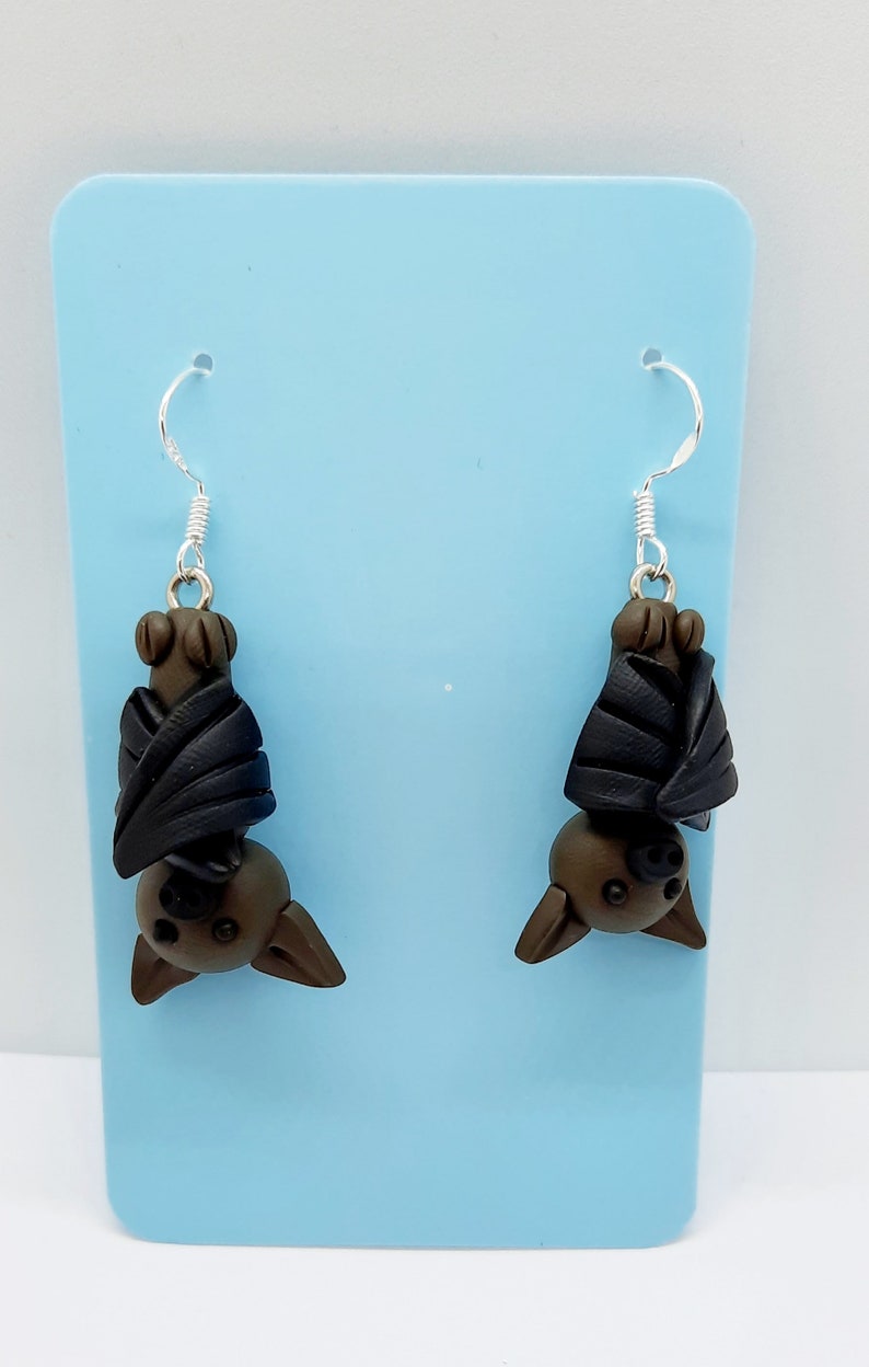 May include: A pair of dangle earrings featuring a brown bat with black wings hanging upside down. The earrings are displayed on a light blue background.
