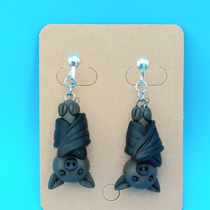 May include: A pair of black bat earrings with silver clip-on backs. The bats are hanging upside down with their wings wrapped around their bodies.