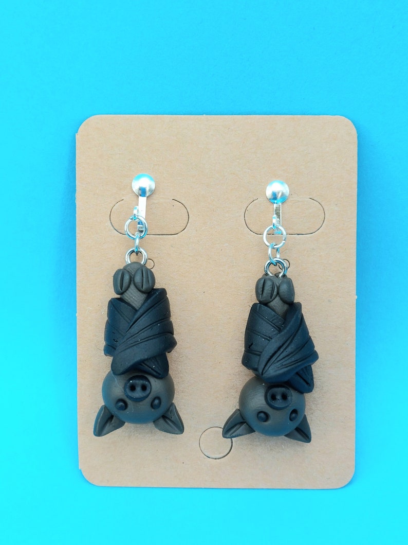 May include: A pair of black bat earrings with silver clip-on backs. The bats are hanging upside down with their wings wrapped around their bodies.