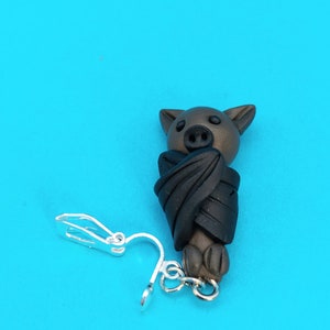 May include: A brown bat with black wings, curled up in a sleeping position, is attached to a silver clip-on earring.