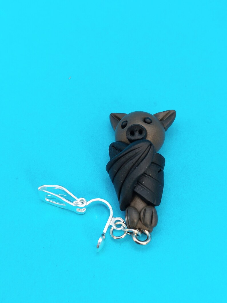 May include: A brown bat with black wings, curled up in a sleeping position, is attached to a silver clip-on earring.