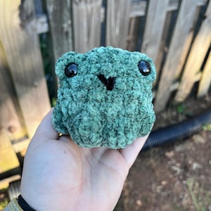 May include: A small, round, green crochet frog plush toy made from textured yarn. The frog features two large, glossy black plastic safety eyes and a small, stitched black V-shaped mouth centered on its face. The surface has a soft, fuzzy appearance.
