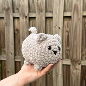 May include: A small, round, light beige crochet plush toy shaped like a cat. The figure features a textured, soft yarn surface, two small pointed ears, a short tail, and two black plastic bead eyes with a small black stitched nose and mouth.