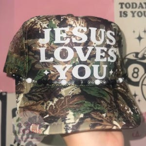 May include: A camouflage print trucker hat featuring the white text JESUS LOVES YOU on the front panel. The hat is accented with a decorative beaded trim across the base of the front panel, consisting of alternating white, black, and silver beads.