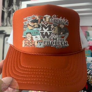 Peut inclure: Une casquette de type trucker orange brûlé avec un filet blanc à l'arrière et un panneau avant en mousse. L'avant présente une impression graphique avec le texte "Raised on Wallen", "Somebody's Problem", "MW", "Wallen 98" et "I'm the Problem", ainsi que des illustrations d'une personne jouant de la guitare.