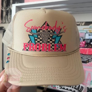 May include: A tan trucker hat with a mesh back and a gold cord trim along the brim. The front features a graphic with a leopard print longhorn skull, two teal lightning bolts, a black and white checkered triangle, and the text Somebody's PROBLEM.