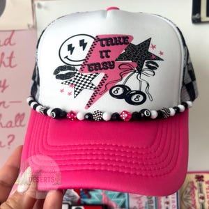 May include: A white and hot pink trucker hat featuring a graphic print of a pink lightning bolt with the text Take It Easy, a smiley face, stars, and billiard balls. The brim is hot pink, and the base is adorned with a string of black and white beads.
