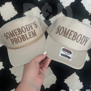 May include: Two beige trucker hats with mesh back panels. One hat says "Somebody's Problem" in brown lettering with a gold chain. The other hat says "Somebody" in brown lettering. Both hats have a small white label with black text.