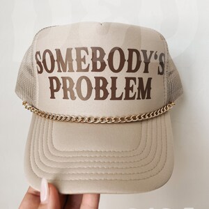 May include: A beige trucker hat with a brown embroidered design that says "Somebody's Problem". The hat has a gold chain around the brim.