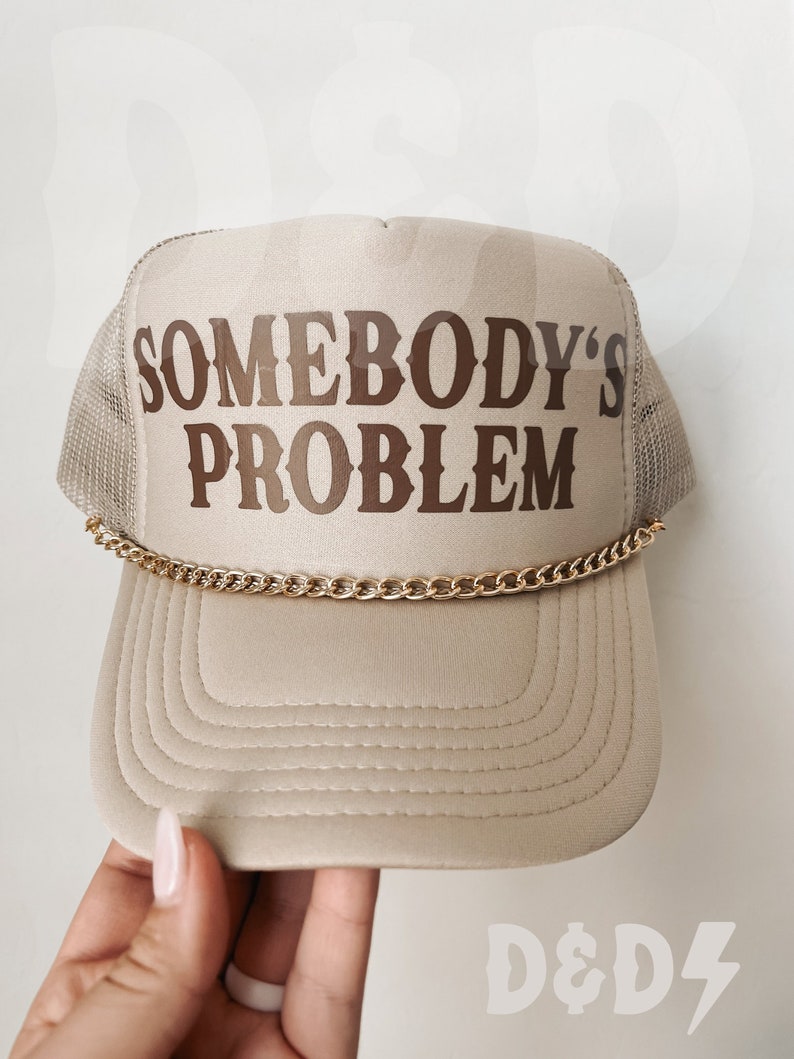 May include: A beige trucker hat with a brown embroidered design that says "Somebody's Problem". The hat has a gold chain around the brim.