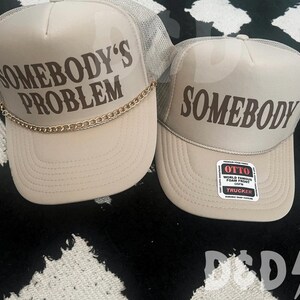 May include: Two beige trucker hats with mesh back panels. The hat on the left has a gold chain around the brim and the text "Somebody's Problem" in brown lettering. The hat on the right has the text "Somebody" in brown lettering. Both hats have a black and white label with the text "Otto World Famous Foam Front OSFM Trucker".
