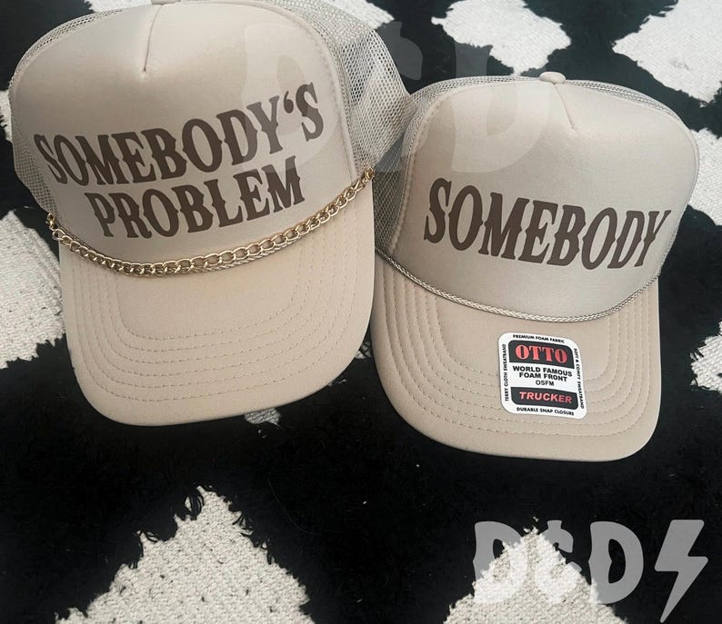 May include: Two beige trucker hats with mesh back panels. The hat on the left has a gold chain around the brim and the text "Somebody's Problem" in brown lettering. The hat on the right has the text "Somebody" in brown lettering. Both hats have a black and white label with the text "Otto World Famous Foam Front OSFM Trucker".