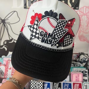 May include: A baseball-themed trucker hat with a white crown, black peak, and red trim. The hat features a baseball graphic with "GAME DAY" text, star and lightning bolt patches, and a chain detail.