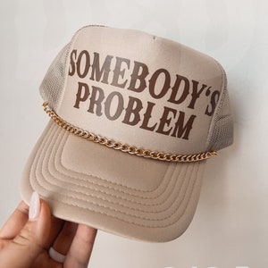 May include: A beige trucker hat with a brown embroidered design that says "Somebody's Problem" and a gold chain attached to the brim.