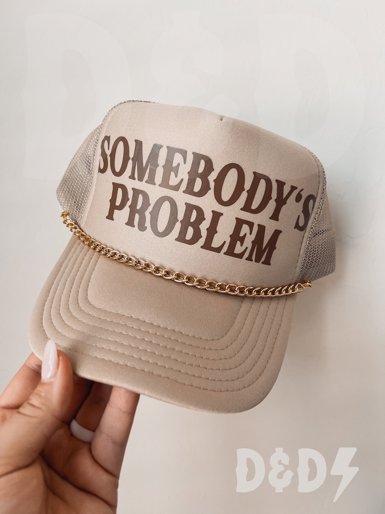 May include: A beige trucker hat with a brown embroidered design that says "Somebody's Problem" and a gold chain attached to the brim.