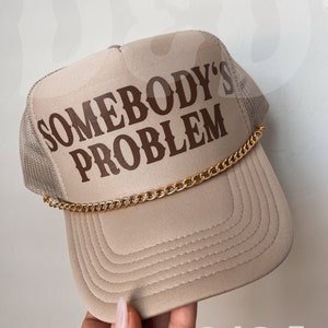May include: A beige trucker hat with a brown embroidered design that says "Somebody's Problem" and a gold chain around the brim. The hat is held by a hand with a white nail polish.