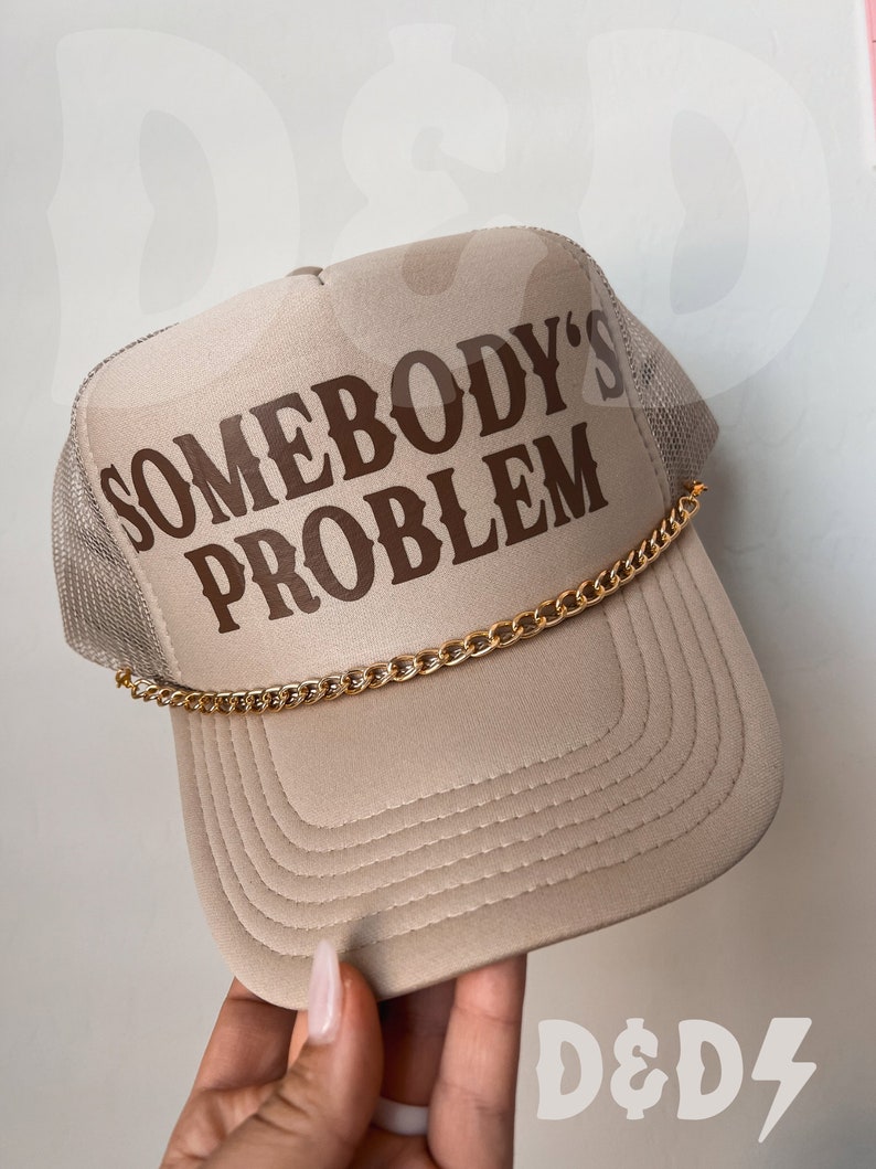 May include: A beige trucker hat with a brown embroidered design that says "Somebody's Problem" and a gold chain around the brim. The hat is held by a hand with a white nail polish.