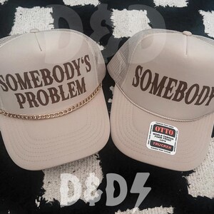 May include: Two beige trucker hats with mesh panels. One hat says "Somebody's Problem" in brown text with a gold chain trim. The other hat says "Somebody" in brown text. The hat on the right has a red and white label that says "Otto World Famous Foam Front Snapback Trucker".