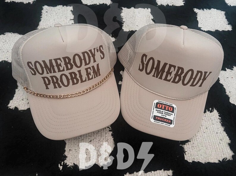 May include: Two beige trucker hats with mesh panels. One hat says "Somebody's Problem" in brown text with a gold chain trim. The other hat says "Somebody" in brown text. The hat on the right has a red and white label that says "Otto World Famous Foam Front Snapback Trucker".