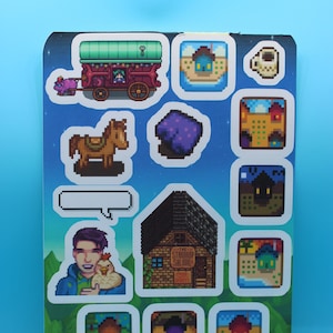 Peut inclure: Une feuille d'autocollants représentant des graphismes pixelisés du jeu vidéo Stardew Valley. Les autocollants comprennent un cheval, un chariot, un tapis violet, une tasse de café, un saloon et divers bâtiments.