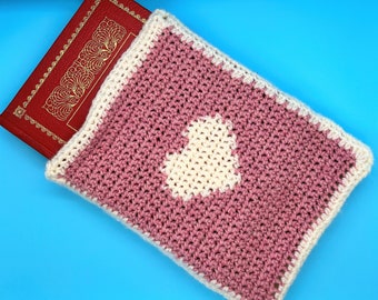 May include: A pink crocheted book cover with a white heart in the centre. The cover is folded over a red book with gold designs.