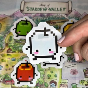 May include: A selection of pixelated Stardew Valley stickers. The stickers depict a white ghost-like character with pink cheeks, a green apple, and a red apple. The background is a map of Stardew Valley, with the title at the top.