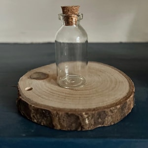 Könnte beinhalten: Eine kleine, klare Glasflasche mit Korken, die auf einer runden Holzscheibe steht. Die Holzscheibe weist sichtbare Jahresringe und einen rauen Rand auf. Die Flasche ist leer und zeigt ihr transparentes Design.