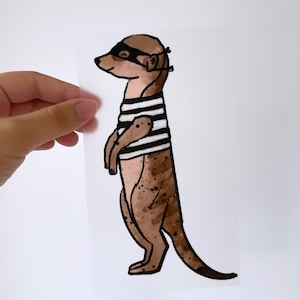 Op de afbeelding: Een bruine en zwarte mangoest met een zwart oogmasker en een gestreept shirt. De mangoest staat op zijn achterpoten met zijn staart gekruld erachter.