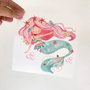 Peut inclure: Une illustration aquarelle d'une sirène aux cheveux roses, une queue verte et des étoiles dorées. Elle est entourée de fleurs et de bulles.