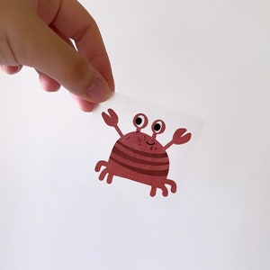 May include: A red cartoon crab with black eyes and a smile. The crab has brown stripes on its body and is holding its claws up.