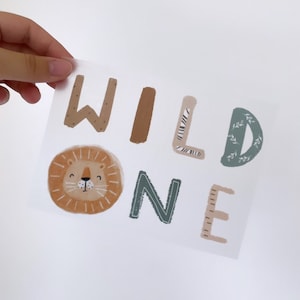 Op de afbeelding: Een lichtbruine, groene en witte sticker met de woorden "WILD ONE" in een speels lettertype. De letter "L" is versierd met een zebraprint, de letter "D" heeft een bladmotief en de letter "O" is een cartoon-leeuwenkop.