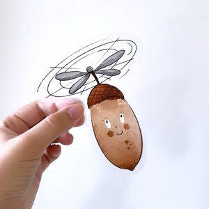 May include: A charming sticker of a cartoon acorn with a propeller. The acorn is light brown with a smiling face and small freckles. The propeller is grey, and the sticker is held by a hand against a white background.