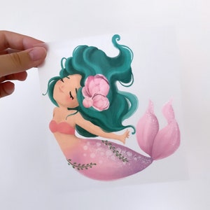 May include: A whimsical illustration of a mermaid with long green hair, a pink tail, and a pink flower in her hair. The mermaid is smiling and has her eyes closed.