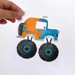 May include: A colourful illustration of a monster truck. The truck has a blue cab and orange top with black tyres and blue and yellow accents. The image is held by a hand.