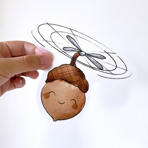 May include: A cartoon acorn with a happy face and a small propeller on top. The propeller is spinning, and the acorn is flying through the air.