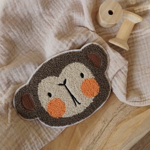 Op de afbeelding: Een bruine en beige pluche aapgezicht applique met oranje geborduurde wangen. De applique is op een lichtbruine stoffen achtergrond.