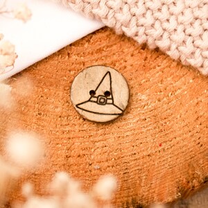 Könnte beinhalten: Ein runder, hellbrauner Knopf mit einem schwarzen Hexenhut-Design. Der Knopf liegt auf einem Holzstück, mit einem hellfarbigen Strickstoff im Hintergrund. Der Knopf ist ein dekoratives Element.