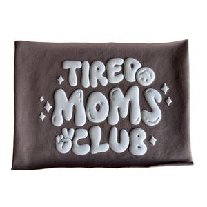 Peut inclure: Un t-shirt marron avec un texte blanc qui dit "TIRED MOMS CLUB" dans une police de caractères amusante et ronde. Le texte est en relief et a un effet 3D.