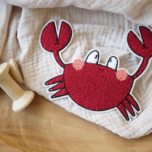 Peut inclure: Applique de crabe en feutre rouge avec des yeux blancs et des joues roses. Le crabe est sur un fond de tissu blanc.