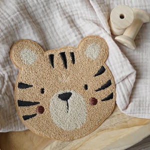Könnte beinhalten: Ein braunes und beiges Plüsch-Tigerkopf-Applik mit schwarzen gestickten Streifen und einem süßen Gesicht. Das Applik befindet sich auf einem hellbeigen Stoffhintergrund.