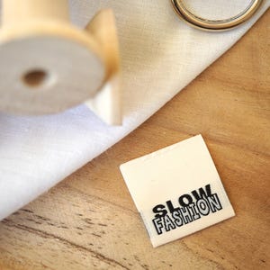 Peut inclure: Une étiquette en tissu blanc avec du texte noir qui dit "SLOW FASHION" sur une surface en bois.