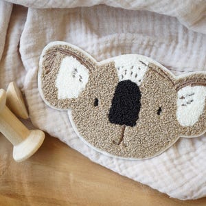 Può includere: Un'applicazione in peluche di koala beige e bianco con occhi e naso ricamati neri. L'applicazione è su uno sfondo di tessuto beige chiaro.