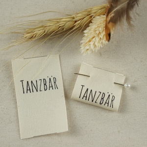 May include: Two white fabric labels with black text that reads "TANZBÄR" on a beige fabric background. One label is pinned to the fabric with a white pearl pin.