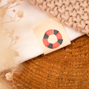 May include: A close-up of a white fabric with a brown fabric label. The label has a red and black circle design with a white background. The fabric is on a wooden surface.