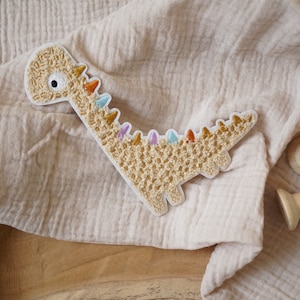 Può includere: Un'applicazione di dinosauro in crochet beige con accenti colorati. L'applicazione è su uno sfondo di tessuto beige.