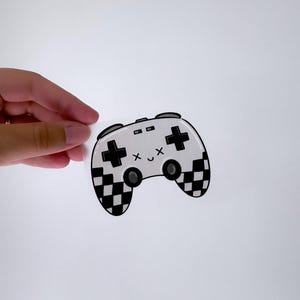 May include: A white and black chequered game controller sticker with a cute cartoon face. The controller has a black outline and black buttons.