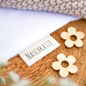 Peut inclure: Une étiquette en tissu blanc avec le texte "NASCHKATZE" imprimé en noir. L'étiquette est sur une surface en bois avec deux petites formes de fleurs en bois.