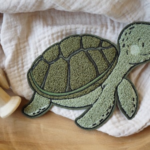 Può includere: Applicazione a forma di tartaruga verde con superficie strutturata e dettagli di cucitura neri. La tartaruga ha un design del guscio dettagliato ed è ambientata su uno sfondo di tessuto neutro. Una bobina di legno è sullo sfondo.