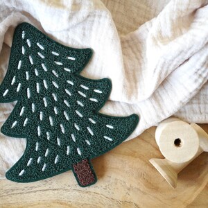Könnte beinhalten: Ein grüner Filz-Weihnachtsbaum-Anhänger mit weißen Nähten. Der Baum liegt auf einer Holzoberfläche mit einem weißen Tuch im Hintergrund.